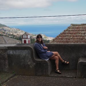 Picture of Emma sat on a stone bench overlooking a view of surrounding villages and the sea.