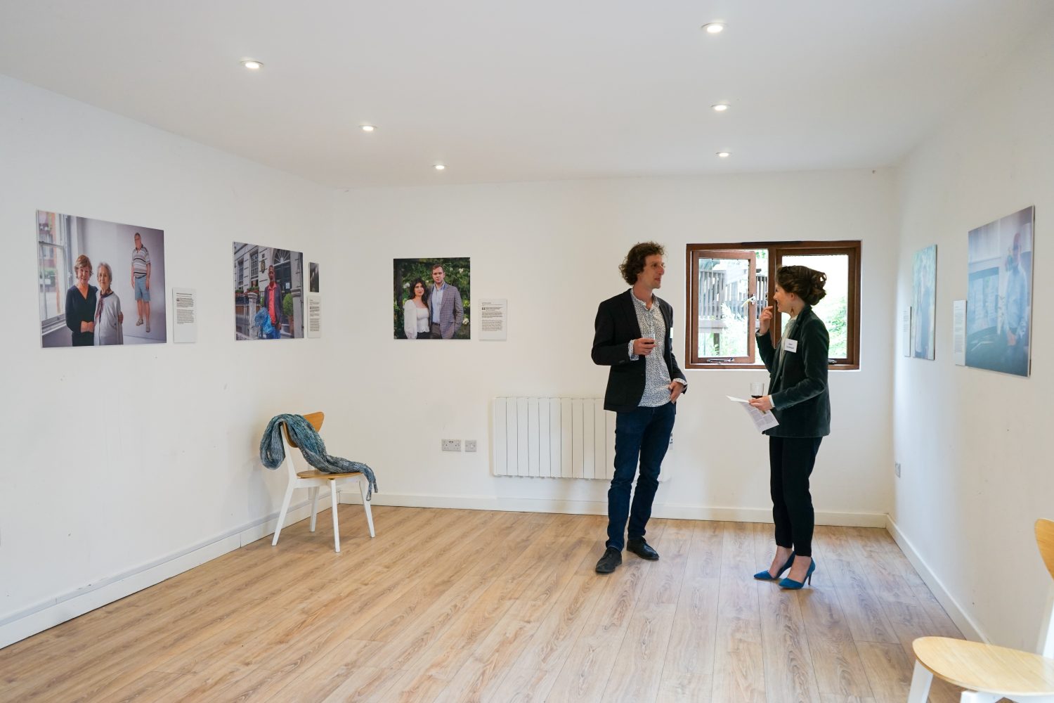 Two people standing talking in the corner of a room with a wooden floor, white walls, and pictures hung up around it.