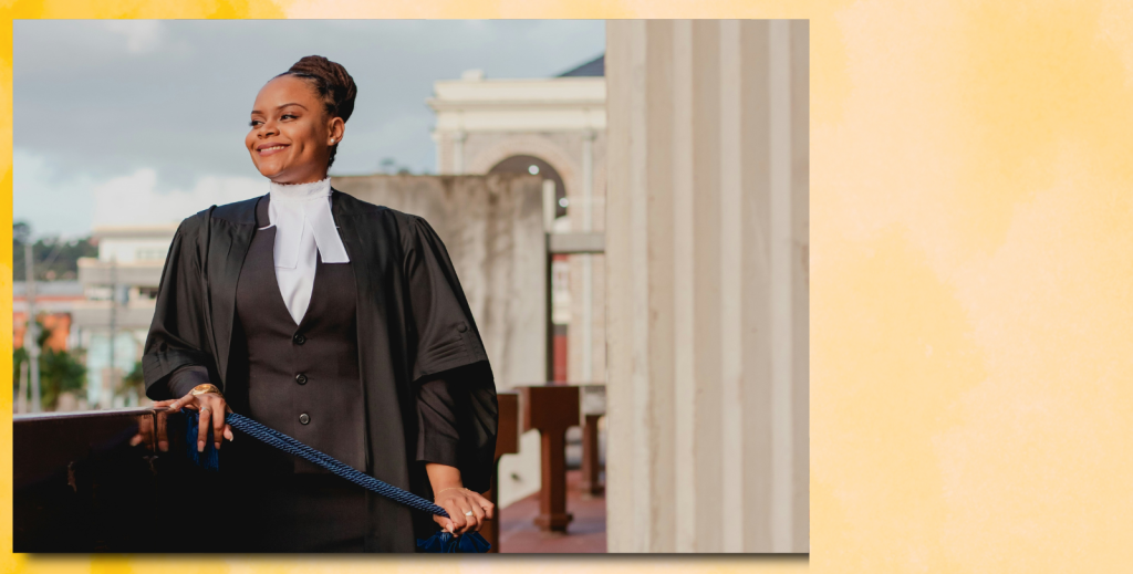 A young Black woman IN law graduate robes stands on a balcony and looks out at the view. She is smiling.